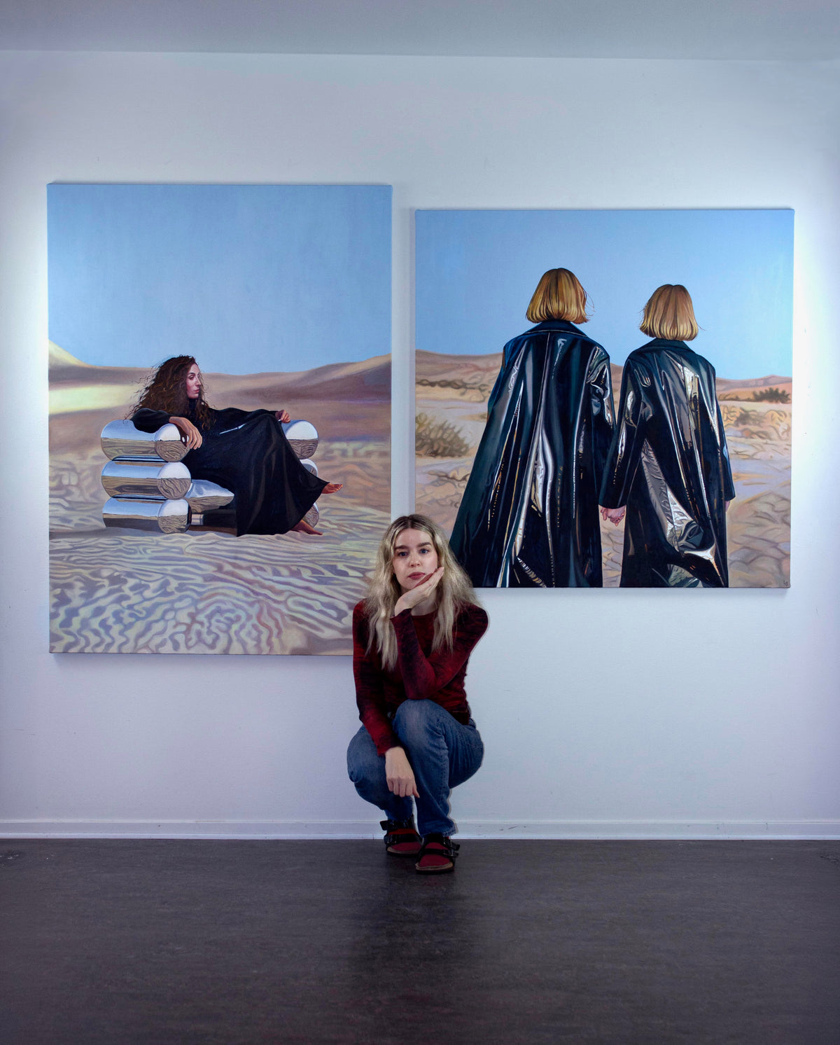 Lovisa Sköld in front of two oil paintings of figures in glossy black coats in a desert